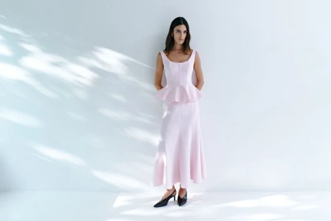 Model wearing a light pink sleeveless peplum top and matching skirt standing against a white wall with soft light patterns.