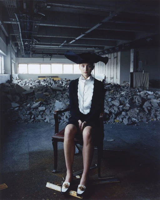 Fashionable woman wearing black dress and oversized hat seated on a chair in an abandoned industrial building surrounded by debris.