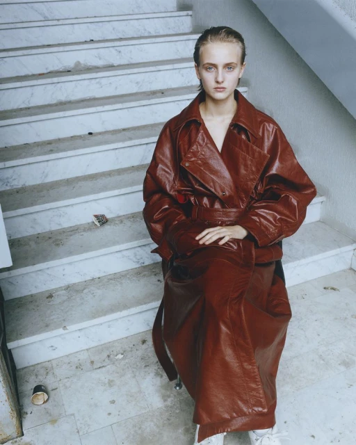 A woman in a long red coat sits on white marble stairs in a minimalist setting, looking at the camera with a neutral expression.