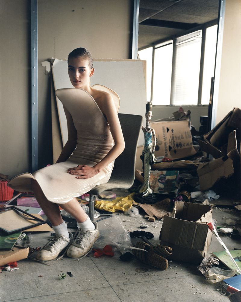 Woman in sculptural dress poses in a messy indoor location with discarded objects. Fashion editorial photography with unique style.