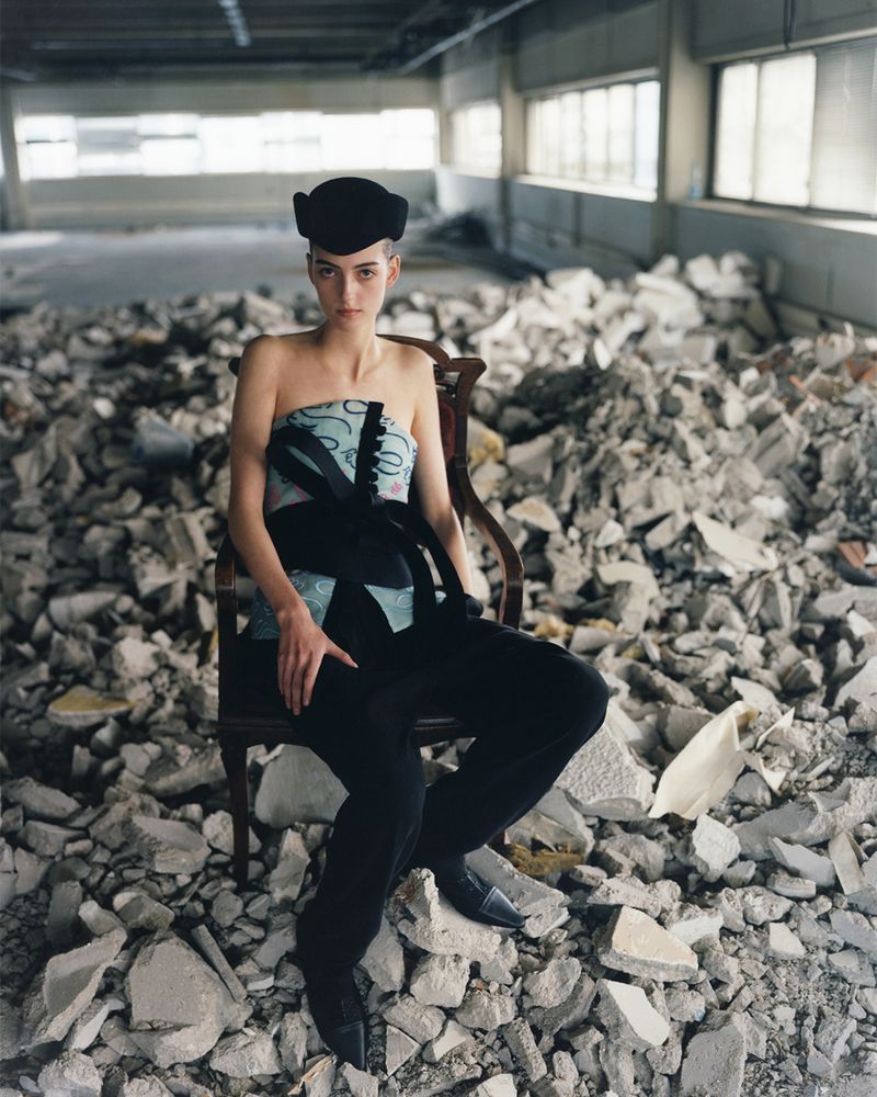 Woman in strapless top and hat sitting in old chair surrounded by rubble in derelict building with large windows and overhead beams.