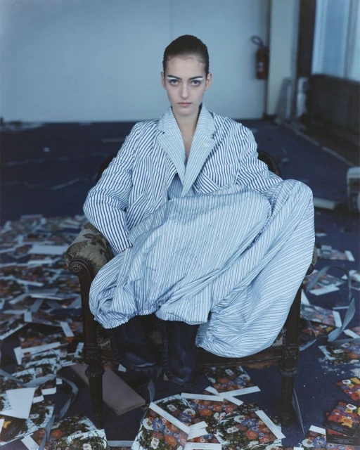 A young woman sits in a chair amidst scattered papers in a room, wearing a striped suit and dark shoes, gazing directly at the camera.