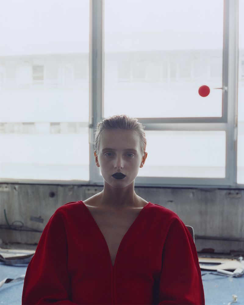 Woman with dark lipstick in a red dress poses in front of a large window with natural light in an interior setting. High fashion concept.