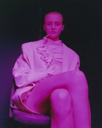 Woman in pink pinstripe blazer poses while seated in an indoor setting with dark backdrop, bathed in pink-purple lighting.