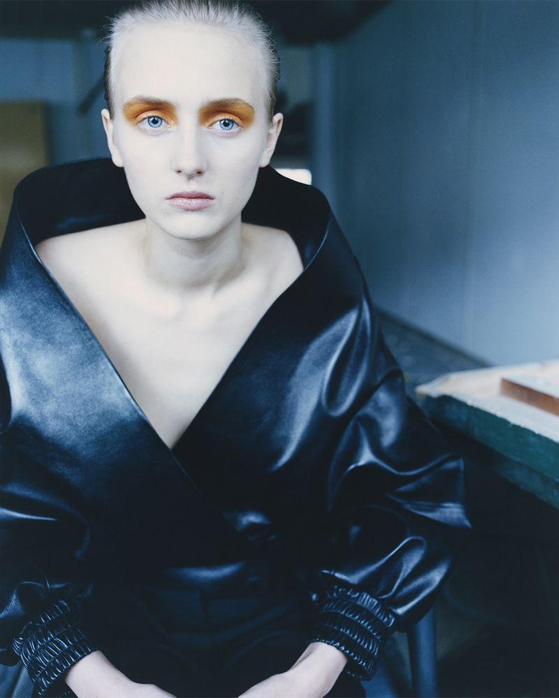 Woman with pale skin and vibrant orange eyeshadow poses in a studio wearing a black, deep V-neck wrap top with gathered sleeves.