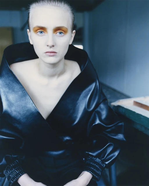 Woman with pale skin and vibrant orange eyeshadow poses in a studio wearing a black, deep V-neck wrap top with gathered sleeves.