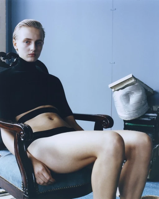 A fair-skinned, blonde-haired person seated in a chair, dressed in a black top and bottoms, poses in front of a blue wall.