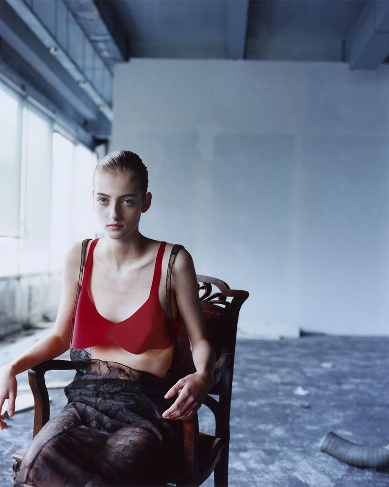 Woman in red top and sheer black skirt sitting in wooden chair in empty industrial space with concrete floors and large window.