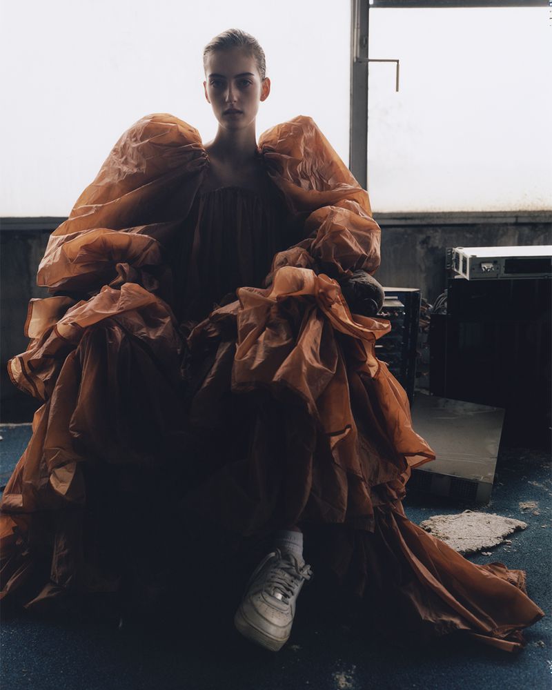 Fashion shot of a young woman in a large copper gown sitting indoors with a window, gray floors, and visible concrete block.