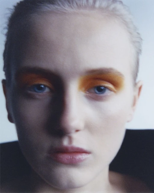 Close-up portrait of a young adult woman with fair skin and blue eyes, featuring striking orange eyeshadow and a dark collar.