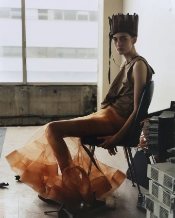 A fair-skinned adult woman with dark hair sits in a chair wearing a layered dress and a cylindrical headdress in an indoor setting.
