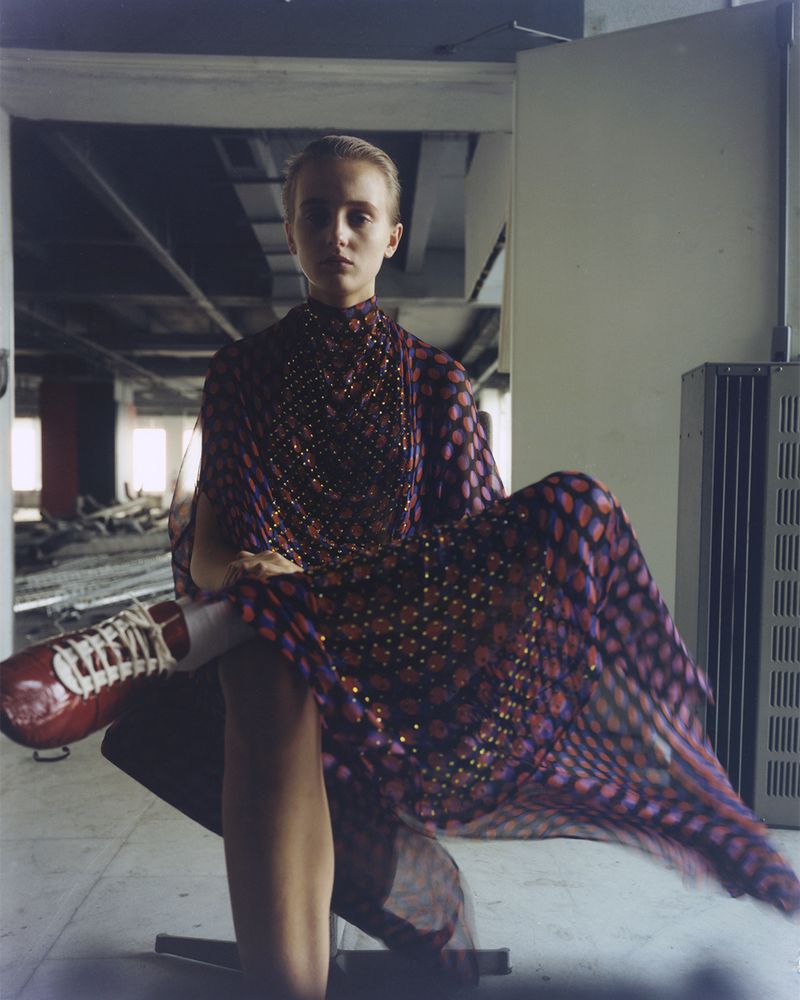 A young woman in a patterned dress sits indoors with red sneakers; the setting is an empty industrial space with natural light.
