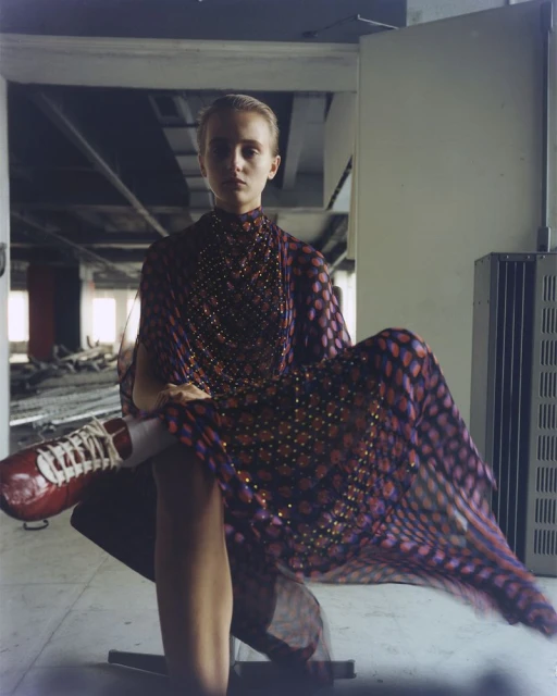 A young woman in a patterned dress sits indoors with red sneakers; the setting is an empty industrial space with natural light.