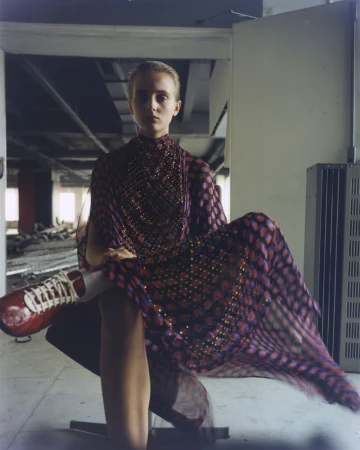 A young woman in a patterned dress sits indoors with red sneakers; the setting is an empty industrial space with natural light.