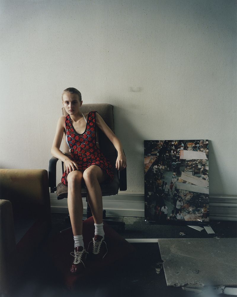 Young woman in a red dress with floral pattern sitting on an office chair in an indoor setting with an abstract art piece in the background.