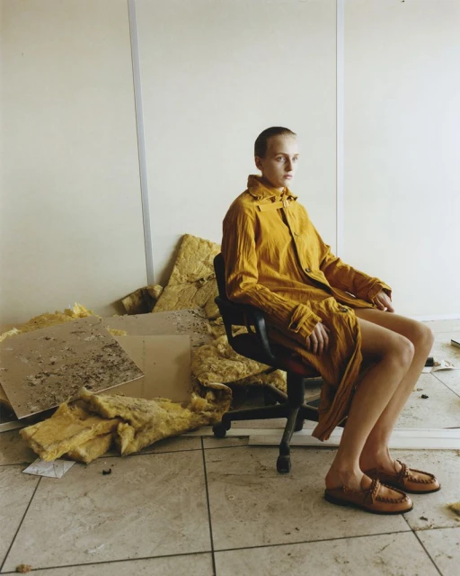 Woman in yellow coat sitting on black chair indoors. Debris surrounds her. Beige walls and tile flooring provide a neutral backdrop.