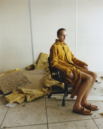 Woman in yellow coat sitting on black chair indoors. Debris surrounds her. Beige walls and tile flooring provide a neutral backdrop.