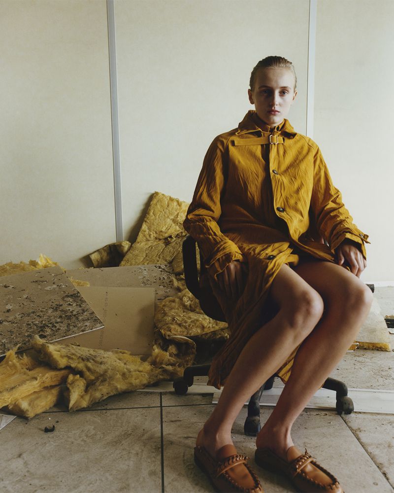 A young woman in a mustard-colored jacket and dress sits in an office chair in a sparse, off-white room amid construction debris.