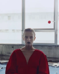 Model with dark lipstick wearing a red V-neck top seated before a large window with a red dot on the glass for D Repubblica No trespassing.