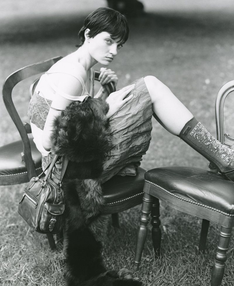 Black and white shot of a woman with short dark hair sitting on chairs outdoors. She wears a dress, socks, and has a purse.