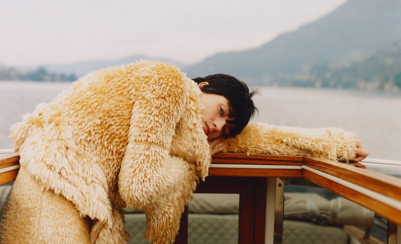 Adult female in a beige coat resting on her arms, overlooking a lake and mountain landscape on an overcast day.