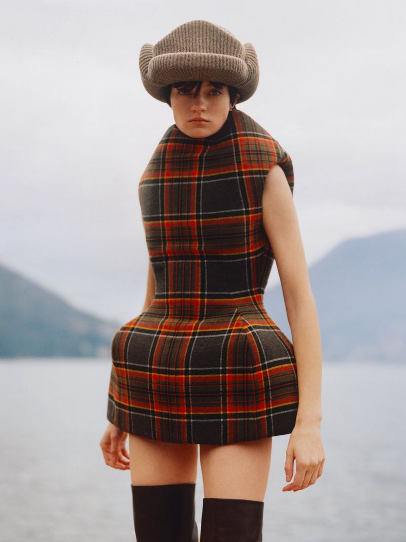 A model wears a plaid dress and trapper hat in an outdoor fashion shot, set against a blurry mountain and lake backdrop.