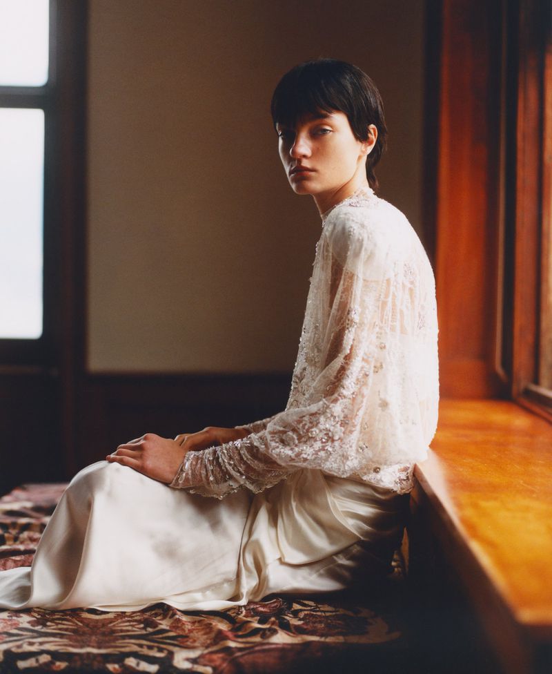 Indoor portrait of a young adult female with short dark hair sitting in a long white dress with lace sleeves, looking toward the camera.
