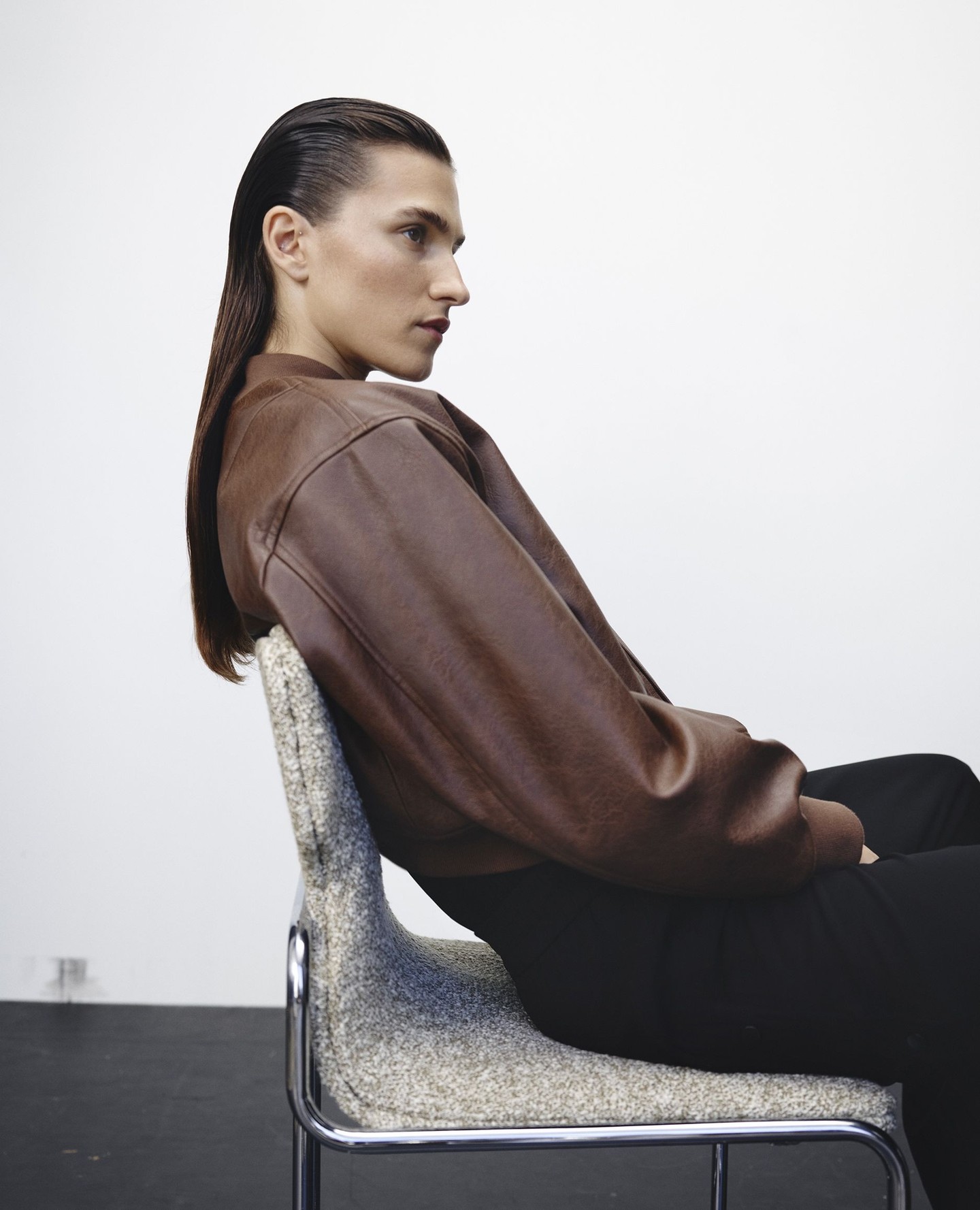 A person with slicked-back hair sits in a chair wearing a brown leather jacket in a studio setting with a white background.