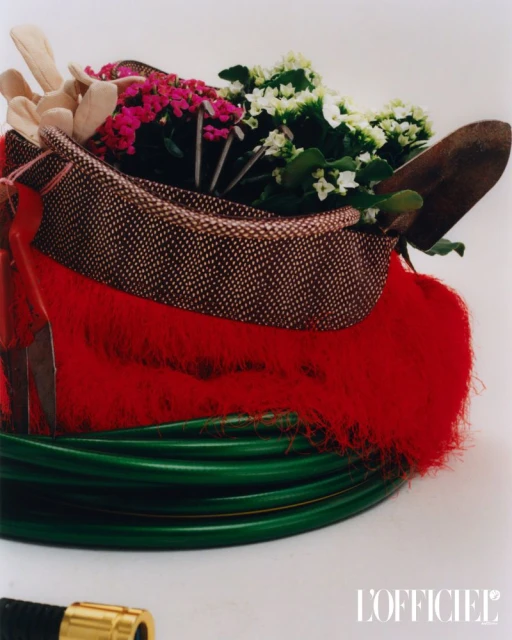Still life featuring a tote bag filled with pink and white flowers, gardening gloves, and a trowel, resting on a green garden hose.