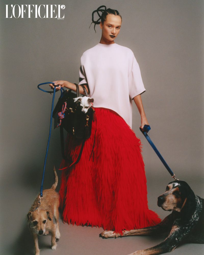Fashion editorial shot of a woman in a white dress and red skirt surrounded by three dogs on leashes in a photo studio.