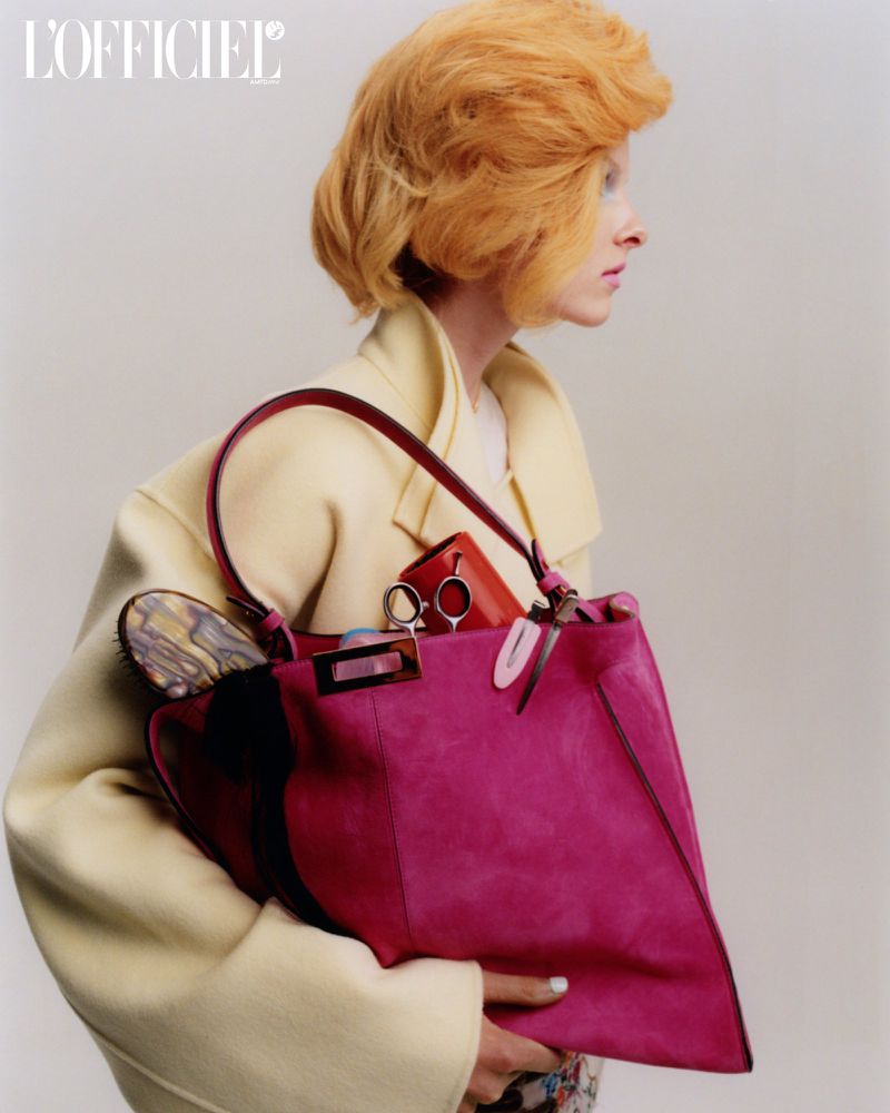 A stylish woman with red hair carrying a pink suede Fendi bag and wearing a cream-colored coat in a fashion studio shot.
