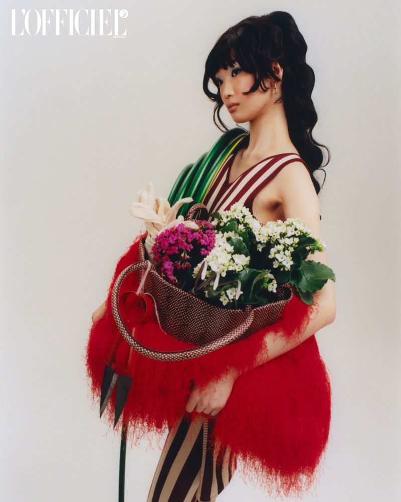 A model poses in a striped dress, holding flowers in a woven basket, draped in a bright red, fringed fabric, against a white background.