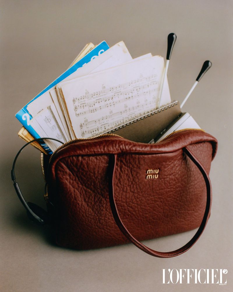 Maroon leather Miu Miu handbag filled with sheet music and conductor's batons set against a neutral background in a studio shot.