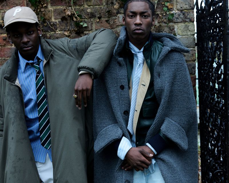 Two Black men pose against a brick wall in stylish overcoats. One wears a baseball cap and the other stands near a dark metal gate.