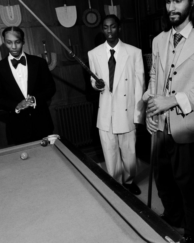 Three men in suits stand around a billiard table in a dimly lit room with decorative paddles on the wall, exuding a vintage vibe.