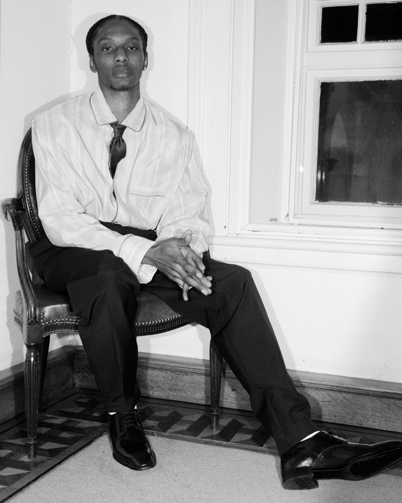 Monochrome portrait of a black man seated in a carved wooden chair wearing a collared shirt, dark tie, and trousers.