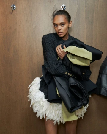 Fashion model in a black boucle jacket and white feathered skirt poses indoors against a wood wall, holding garments in her arms.