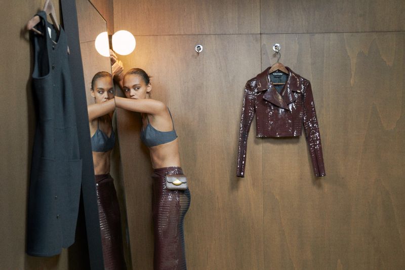 Fashion shot of a slim woman in a bra top and skirt leaning against a mirror in a wooden paneled dressing room, with a jacket on the wall.