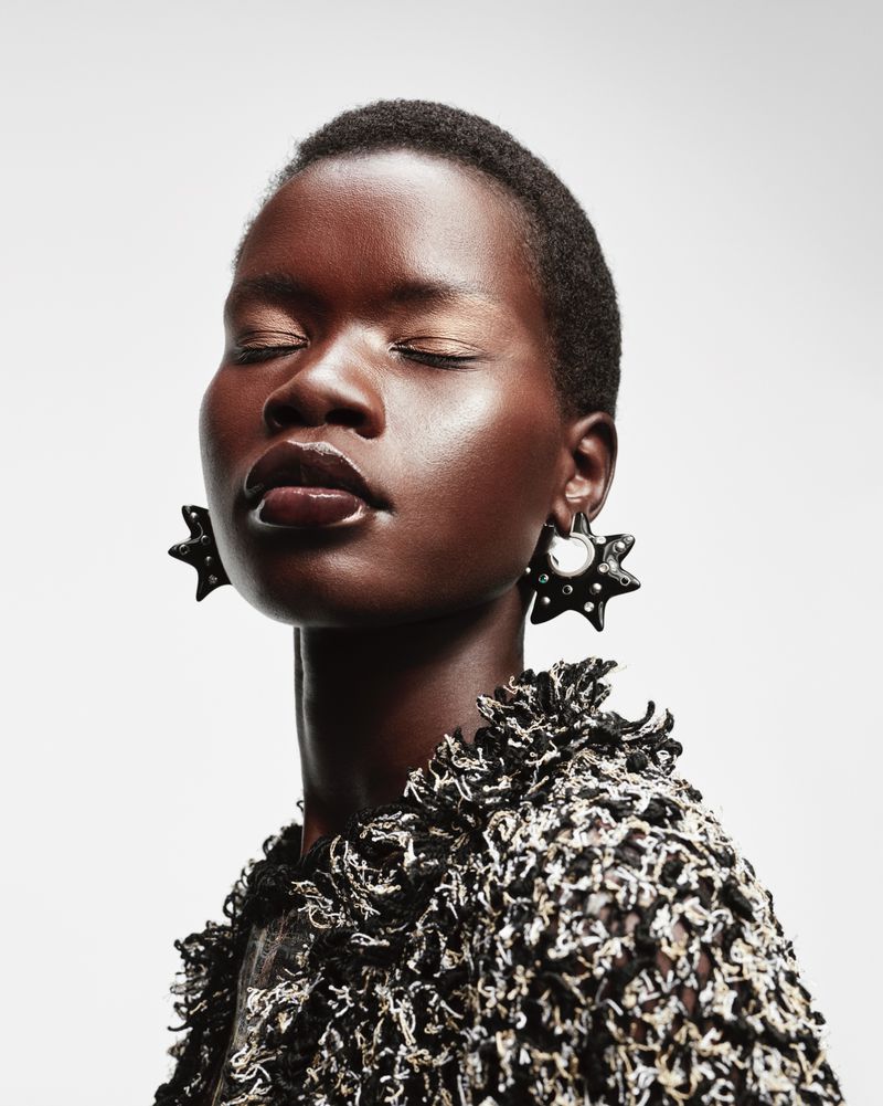 Close-up portrait of a dark-skinned woman with closed eyes, wearing large black starburst earrings and a textured black and white jacket.