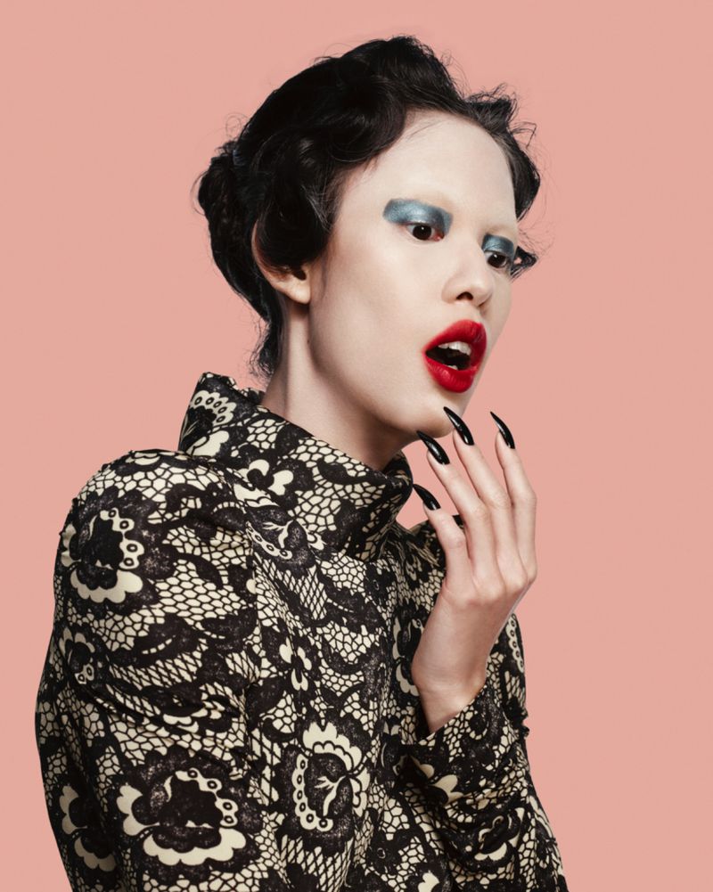 Eye-level portrait of a woman wearing graphic black and beige jacket, bold makeup, long black nails and black hair on pink backdrop.