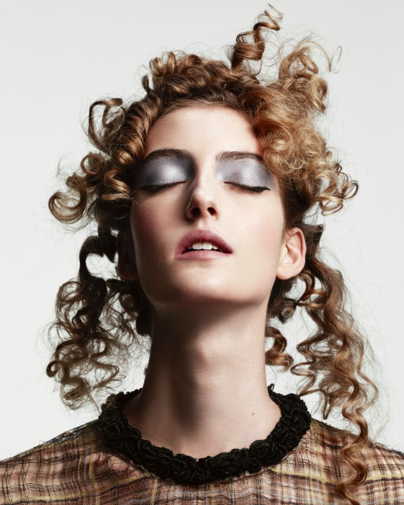 Close-up of a stylish adult woman with curly hair and silver eyeshadow posing against a light background in a fashion portrait.
