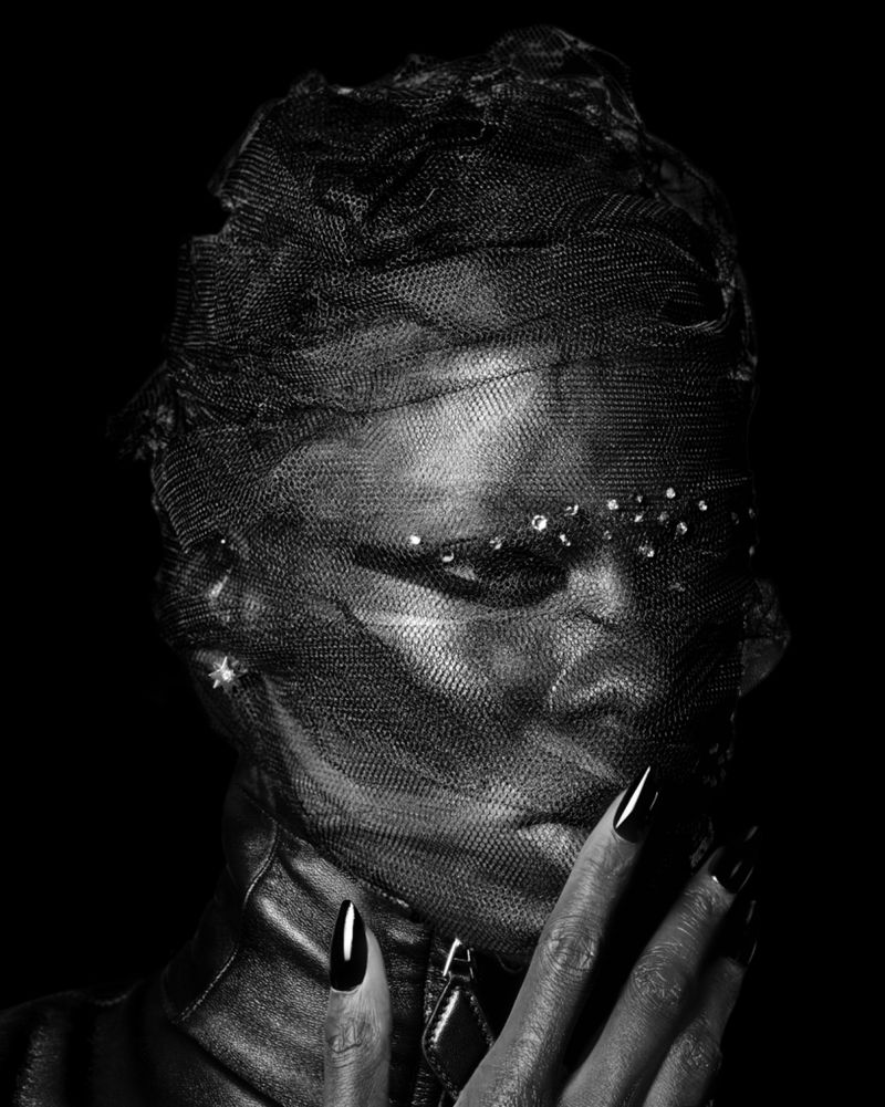 Monochromatic portrait of an adult woman with black nails and a black veil with gem detailing, posing in front of a dark background.