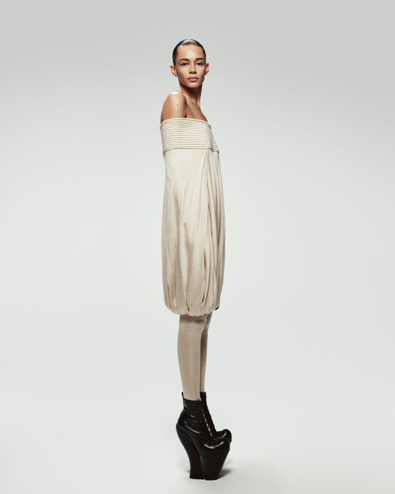 A female model poses in a beige strapless dress with black platform boots against a white background in a fashion forward studio shot.
