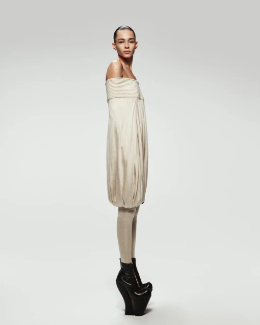 A female model poses in a beige strapless dress with black platform boots against a white background in a fashion forward studio shot.