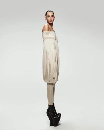 A female model poses in a beige strapless dress with black platform boots against a white background in a fashion forward studio shot.