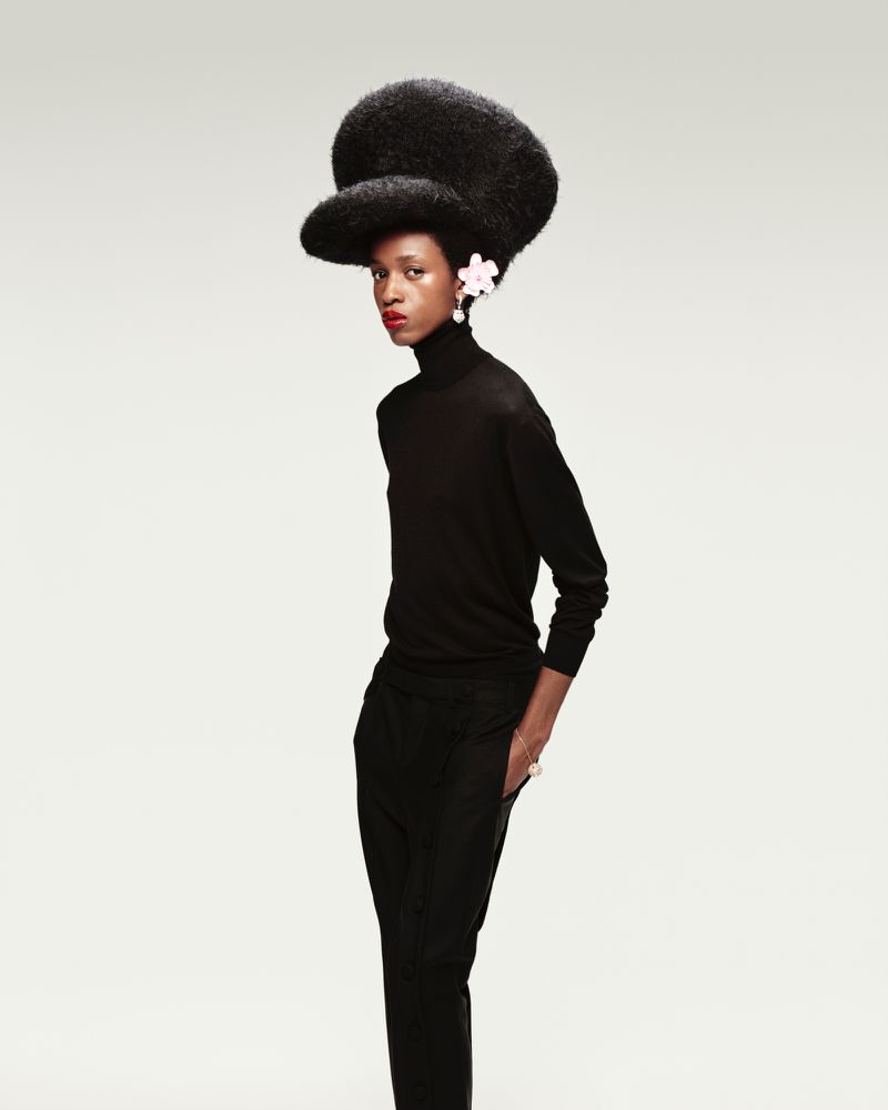 Elegant black woman wearing a black turtleneck and pants with a large Afro hairstyle with white floral details in front of a neutral background.