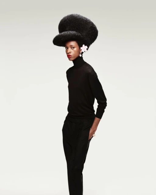 Elegant black woman wearing a black turtleneck and pants with a large Afro hairstyle with white floral details in front of a neutral background.