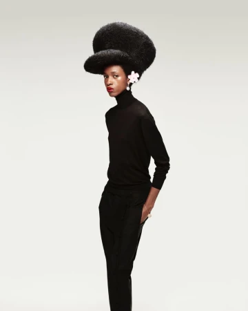 Elegant black woman wearing a black turtleneck and pants with a large Afro hairstyle with white floral details in front of a neutral background.