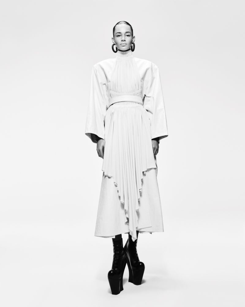 Black-and-white studio shot of a woman in a white dress with a pleated skirt, long sleeves, large earrings, and black platform boots.