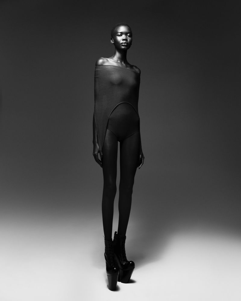 Striking full-length studio shot of a bald, slender Black woman in a draped garment and platform heels, posing against a gradient gray background.